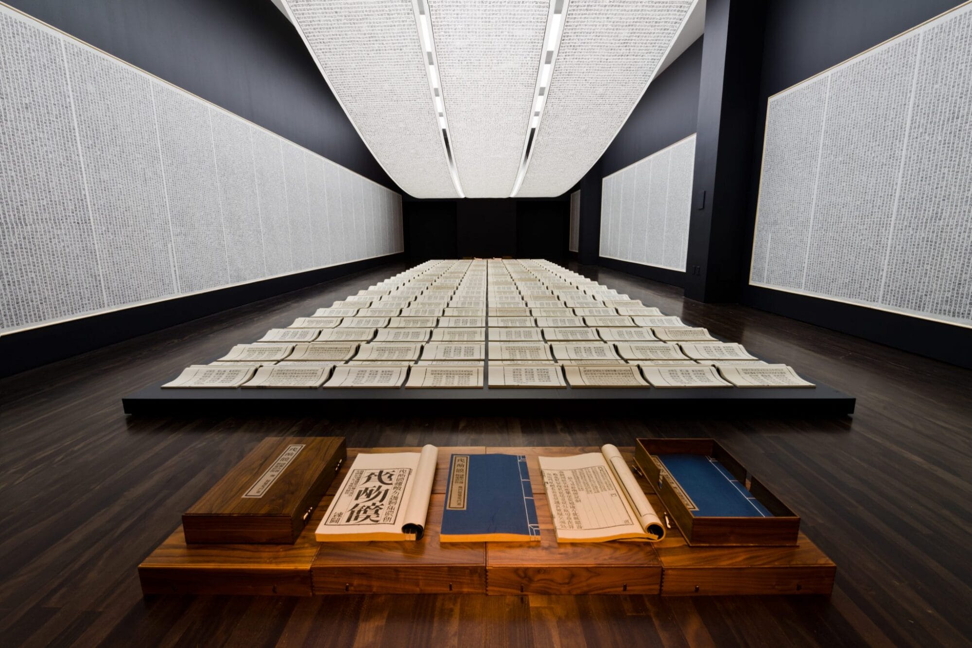 A long, narrow room with large sheets of paper with Chinese writing posted on the wall and draped from the ceiling. Smaller pages lay in a grid in the middle of the floor.