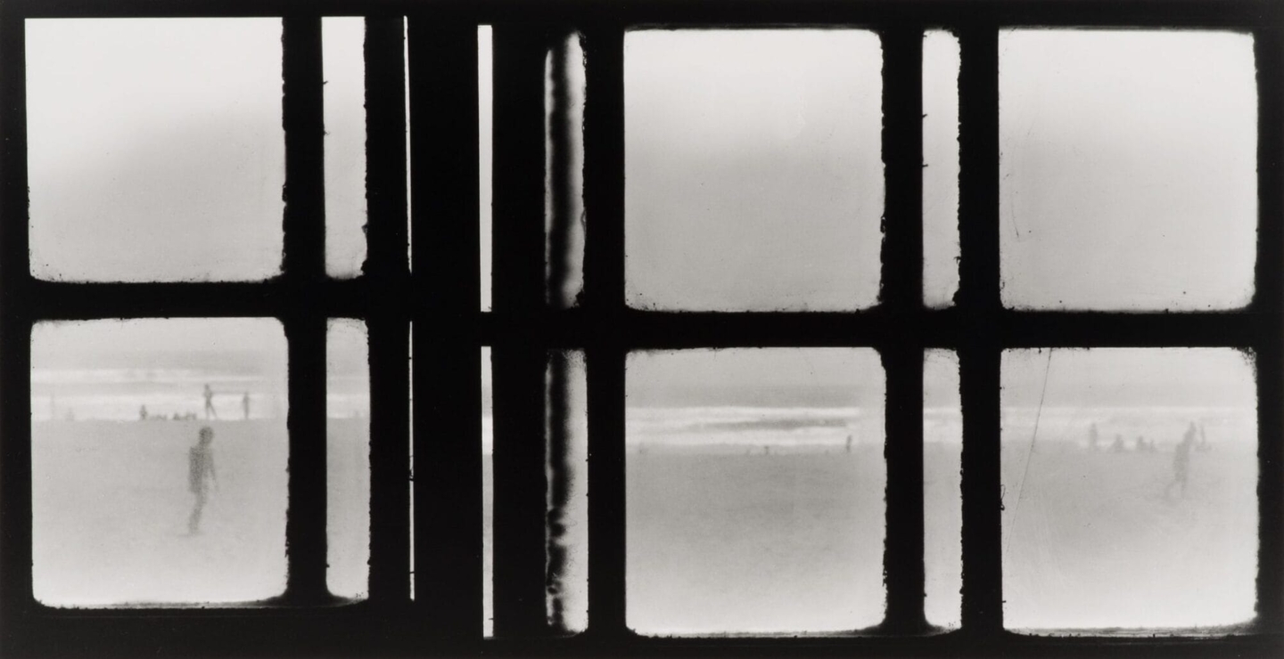 Silhouette of overlapping square window panes. Beyond the window are people walking around on a beach with water in the background.