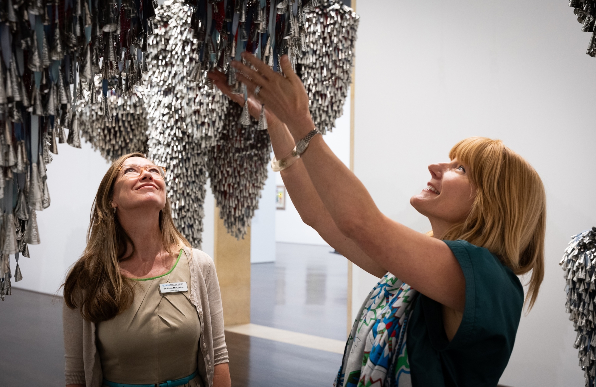 A woman clutching a dangling artwork covered in silver jingles, typically used in Native American dance rituals. A museum educator stands beside her smiling