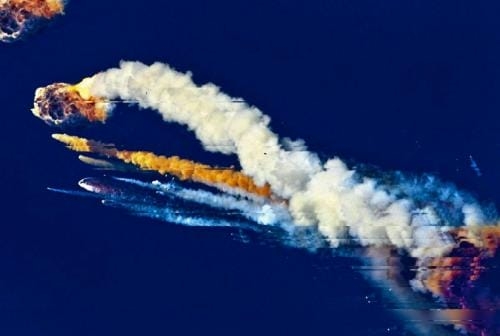 A rocket mid-air disintegration, with trails of smoke and flames arching across a deep blue sky, marking its breakup path.