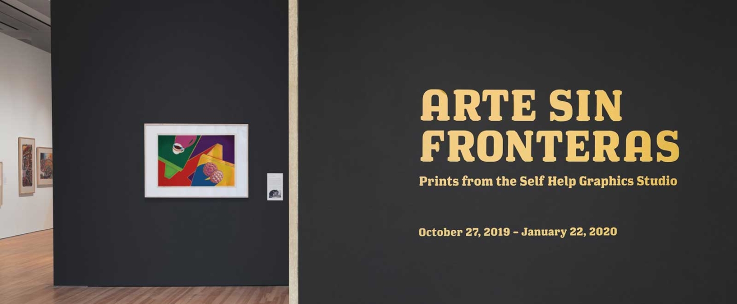 An exhibition wall with the title 'Arte Sin Fronteras: Prints from the Self Help Graphics Studio' displayed prominently in large, bold, yellow text. The exhibition dates are listed as October 27, 2019 - January 22, 2020. To the left, there is a framed colorful abstract artwork on a dark wall, with additional framed artworks visible in the background on a lighter wall.