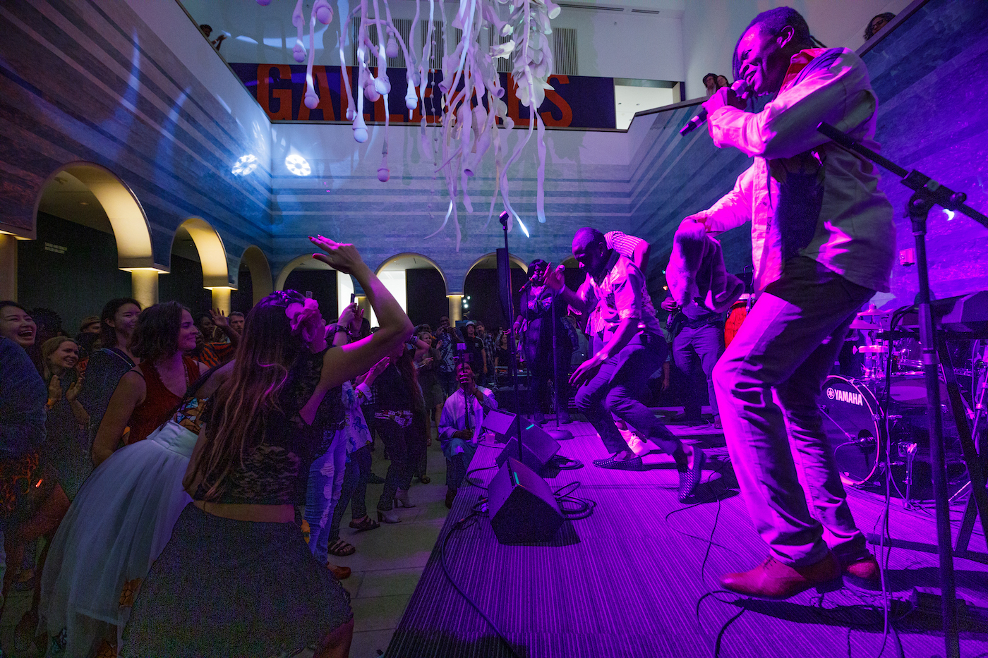Musicians perform on stage in the Blanton atrium while people dance. It is nightime and purple lights color the stage.