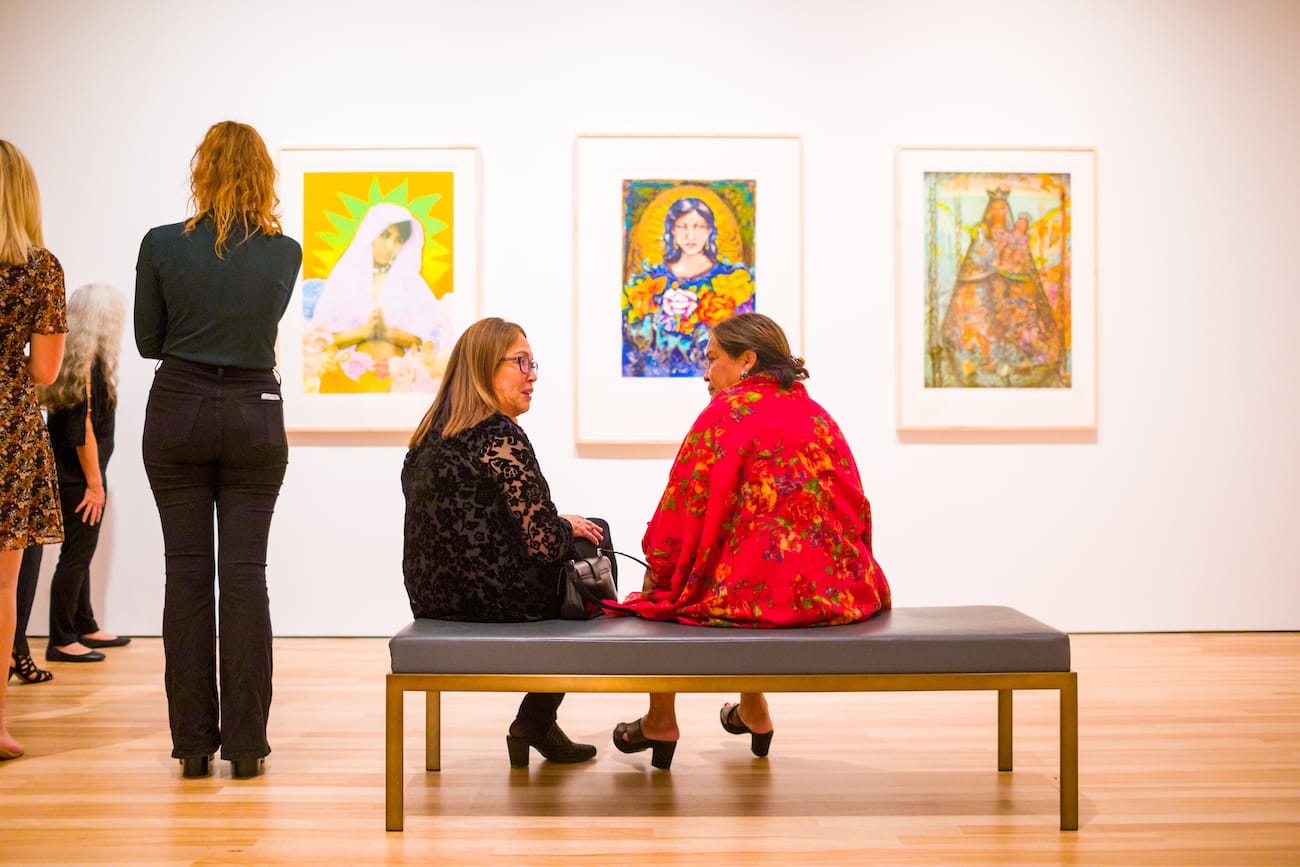 Vibrantly dressed older women sitting on a bench in front of colorful artworks having a conversation. In the background other visitors admire art.
