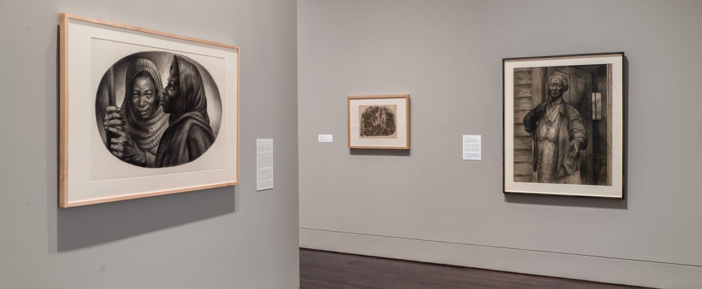 An art gallery featuring three framed artworks on display. The left artwork shows a person in a hooded garment holding a staff, depicted in a circular frame. The center artwork is smaller, with intricate, abstract patterns. The right artwork portrays a person standing in front of a wooden structure, wearing a coat and holding an object. Each piece has a descriptive label next to it, highlighting different styles and subjects of art