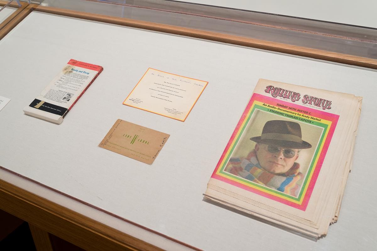 Back cover of a book, a copy of Rolling Stone magazine and two printed cards with some handwritten notes in a display case.