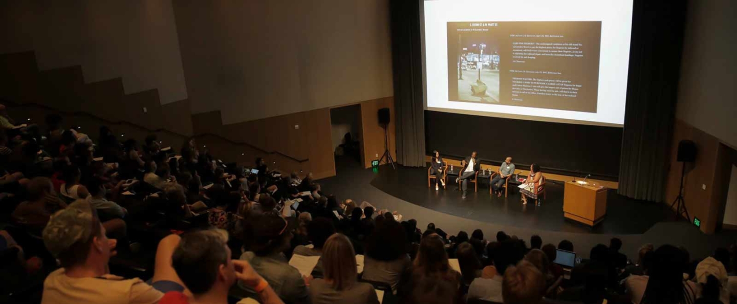 A large audience seated in an auditorium, attentively watching a panel discussion on stage. Four panelists are seated on chairs, with a large screen behind them displaying a slide titled 'I. LOOK AT A PHOTO.' The slide includes a photo of a street scene and accompanying text. The auditorium is dimly lit, with the focus on the stage and screen.