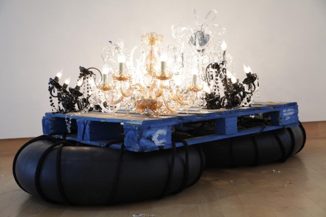 An artistic installation featuring a blue wooden pallet placed on top of large black inner tubes. On the pallet, there are multiple ornate chandeliers with lit bulbs of various designs and colors, including clear, black, and gold. The combination of industrial materials (pallet and inner tubes) with the elegant chandeliers creates a striking contrast, making the installation visually interesting and thought-provoking.