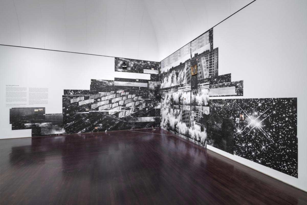 An art installation in a gallery space consisting of a large-scale collage that covers two adjacent walls. The collage features a mix of black-and-white photographs and images of urban landscapes, buildings, and starry skies, arranged in a fragmented, overlapping manner. The dark, reflective floor adds to the installation's dynamic and somewhat chaotic visual effect. The left wall includes a block of text providing context or information about the artwork.