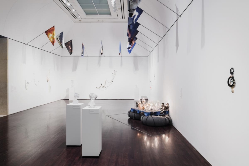 An art gallery featuring various contemporary art installations. The room has white walls and a dark wooden floor. Several pieces of art are displayed, including sculptures on pedestals, wall-mounted artworks, and hanging installations. Colorful triangular flags hang from the ceiling, and a large installation on the floor appears to be made of various materials and lights. The space is well-lit with overhead lighting, and the artworks are arranged to allow viewers to walk around and observe them from different angles.