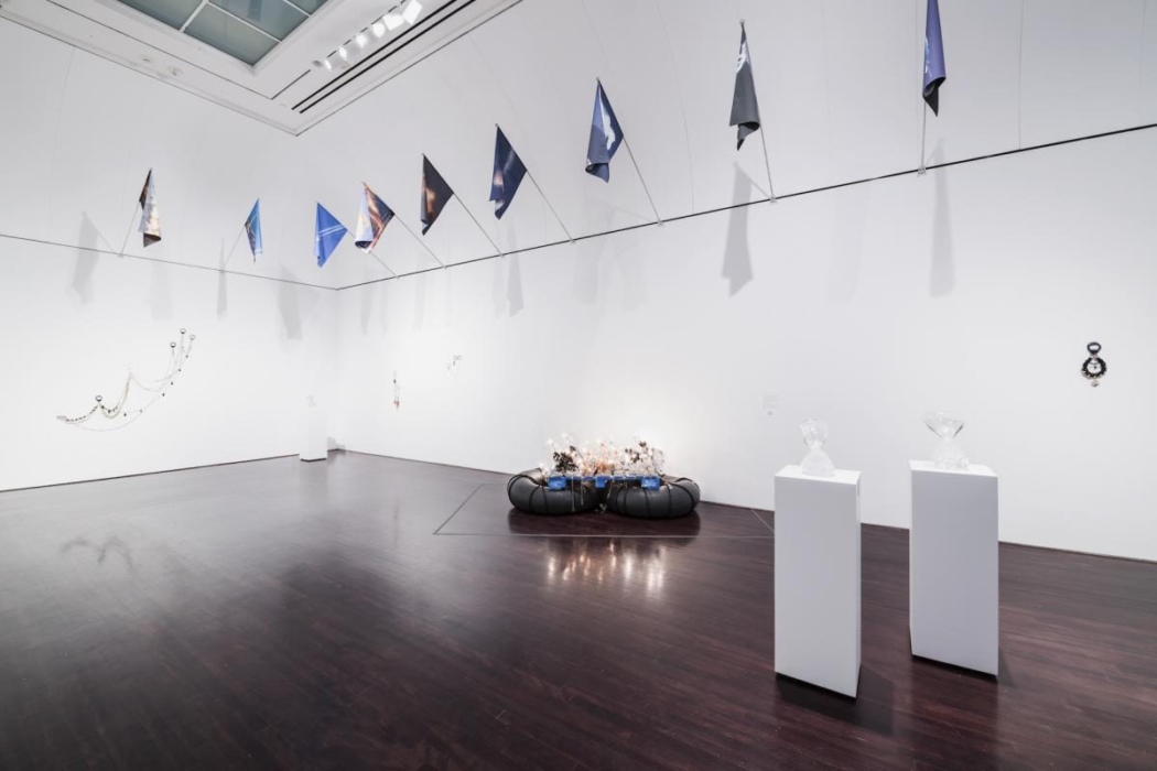 An art gallery or exhibition space with various contemporary art installations. The room has white walls and a dark wooden floor. Several pieces of art are displayed, including: A series of triangular flags hanging from the ceiling. A wall-mounted installation with chains and spherical objects on the left wall. A circular installation on the floor with what appears to be lights or reflective materials. Two white pedestals with transparent sculptures on top. A small wall-mounted piece on the right wall. The overall atmosphere is modern and inviting, encouraging viewers to explore and appreciate the diverse artworks on display.