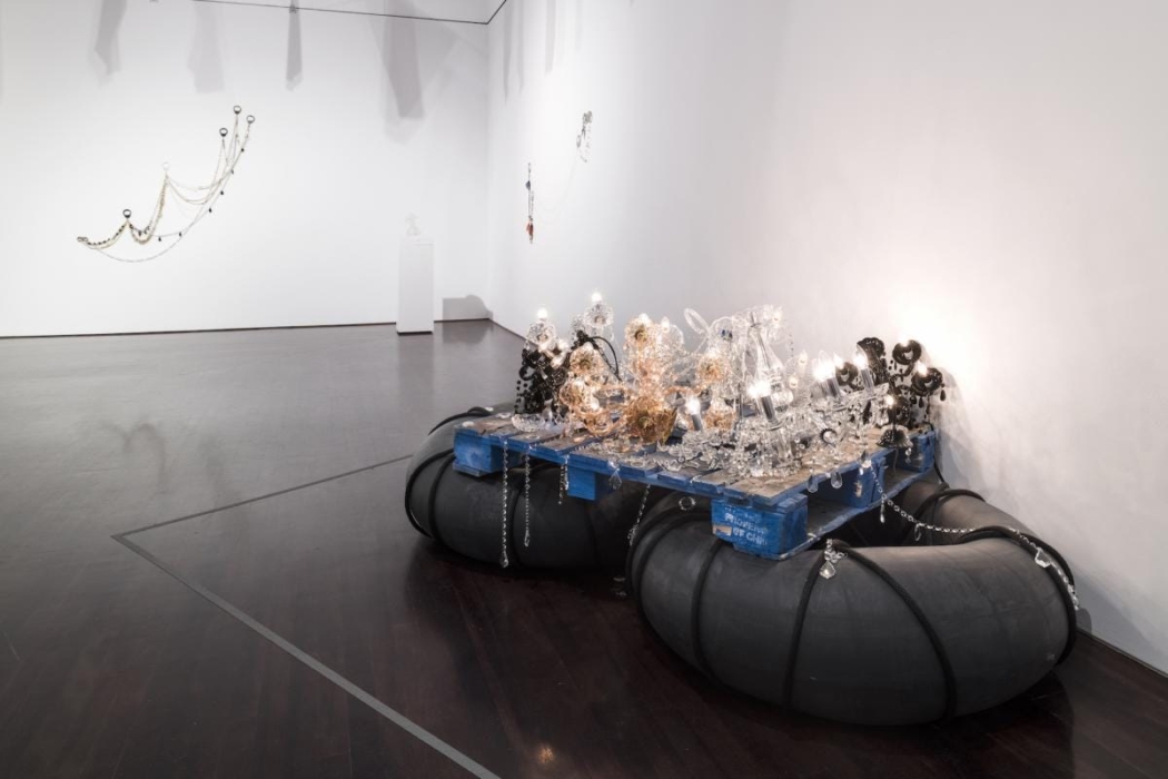 An art installation in a gallery space features a blue wooden pallet supported by large black inflatable tubes. Intricate glass sculptures and lit light bulbs are placed on the pallet. The sculptures are made of clear and colored glass, with some resembling chandeliers. In the background, there is a wall-mounted installation of hanging chains and spherical objects. The gallery has a polished dark floor and white walls, with spotlights illuminating the artworks.