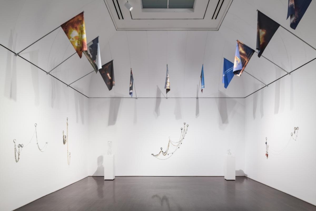 The image shows an art gallery with various installations. The installations include hanging triangular flags with different designs and colors suspended from the ceiling. On the walls, there are several chain-like sculptures made of various materials, including beads and metallic elements, arranged in artistic patterns. Additionally, there are two white pedestals, each displaying a transparent sculptural piece. The gallery has a minimalist design with white walls and a dark floor, creating a clean and modern aesthetic that highlights the artwork. This setting is interesting as it showcases contemporary art in a thoughtfully curated space, emphasizing the interplay between different forms and materials.