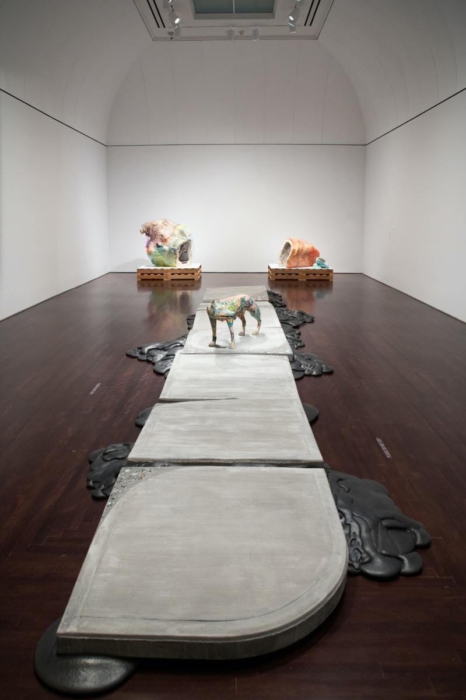 The image shows an art installation in a gallery space. The installation features a pathway made of large, flat, concrete slabs with irregular black shapes surrounding it, resembling melted material. At the center of the pathway, there is a colorful sculpture of an animal, possibly a dog. In the background, there are two large, abstract, colorful sculptures placed on wooden pallets. The gallery has white walls and a high, arched ceiling with spotlights illuminating the artwork. The installation is interesting due to its combination of industrial materials and organic forms, creating a contrast between the rigid and fluid elements.