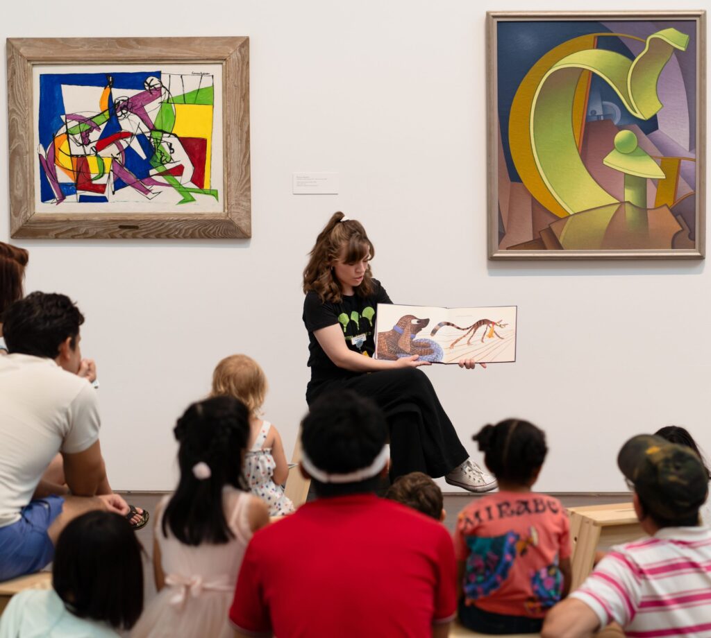 Storytime program. Person sitting in front of framed works of art on the wall of a gallery holding up a children's book. Children and adults sit on the ground facing the person reading.