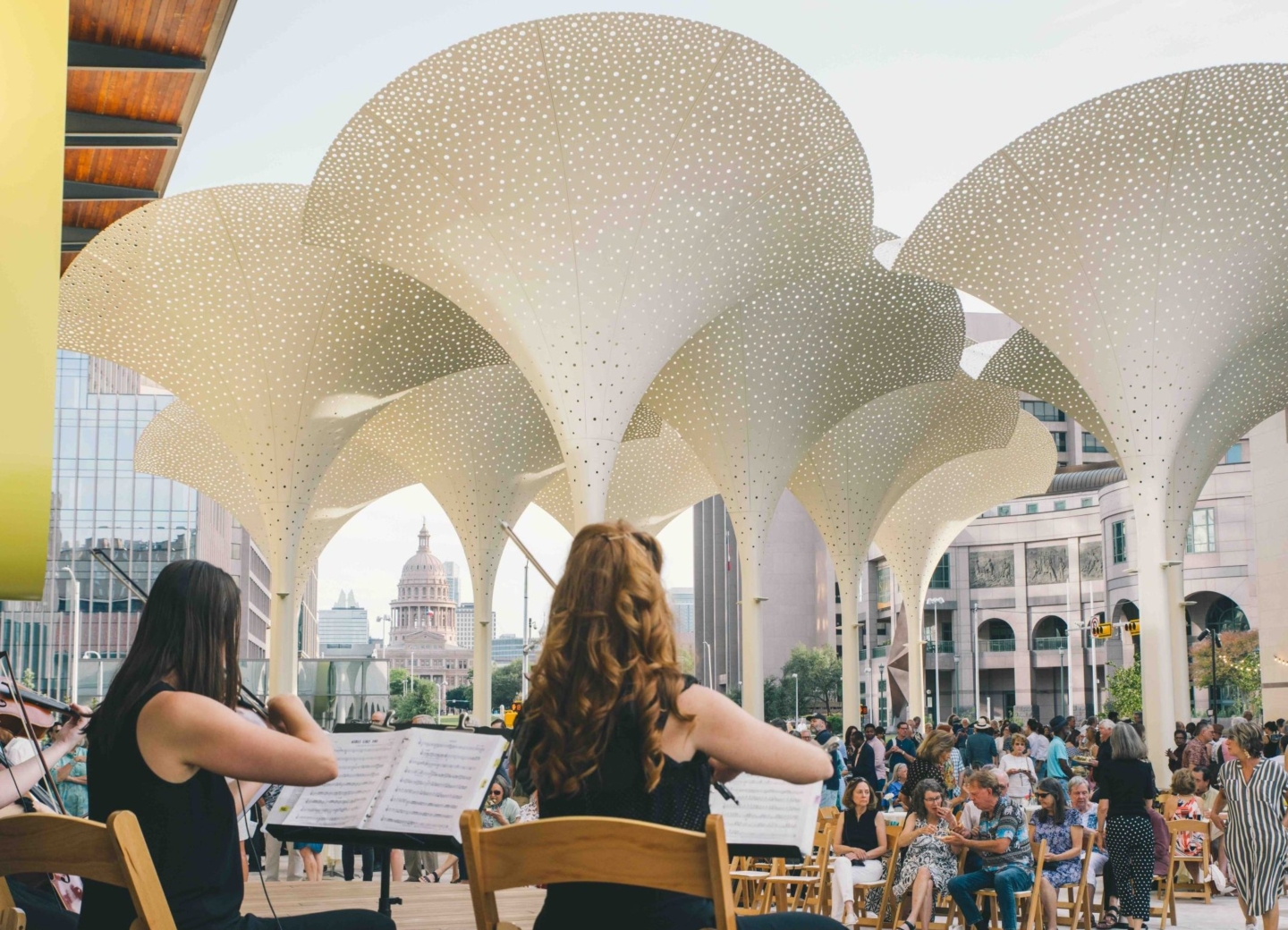 String quartet playing on the grounds of the Blanton Museum of Art to a crowd of seated people. In the background are large petal-like structures and the Texas Capitol building.