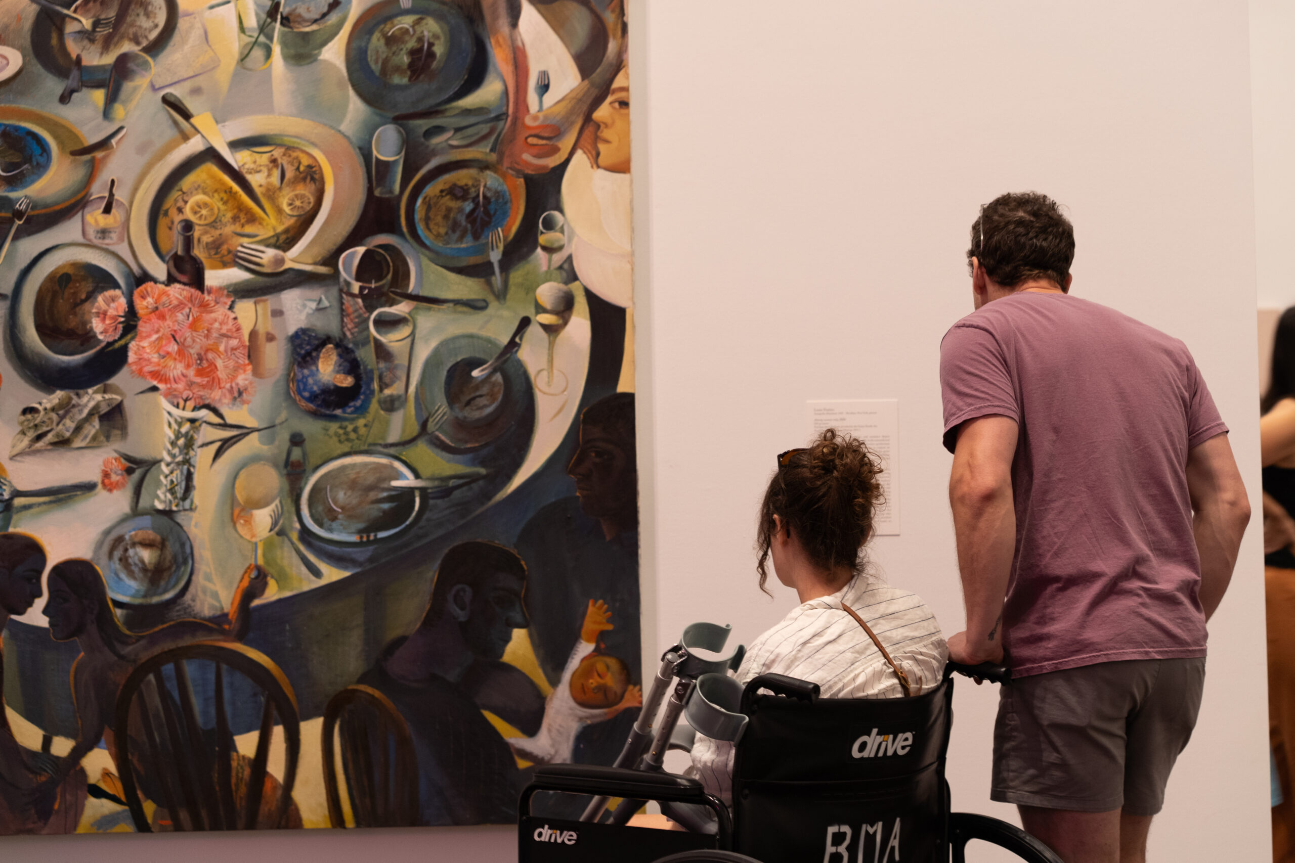 Women in a wheelchair with crutches looking at a colorful painting of people around a table. A man stands next to her reading the artwork label.