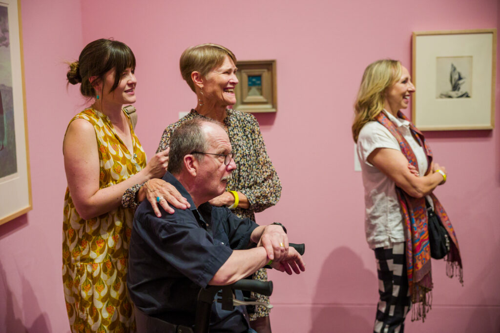 Group of four people stand in a gallery space with framed artwork and pink walls. 