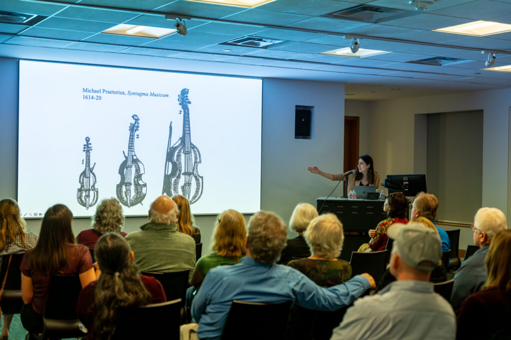 Person stands at front of room gesturing toward a presentation screen that depicts drawings of three upright base instruments with the text "Michael Praetorius, Syntagma Musicum 1614-20" while people sit in chairs looking toward the speaker