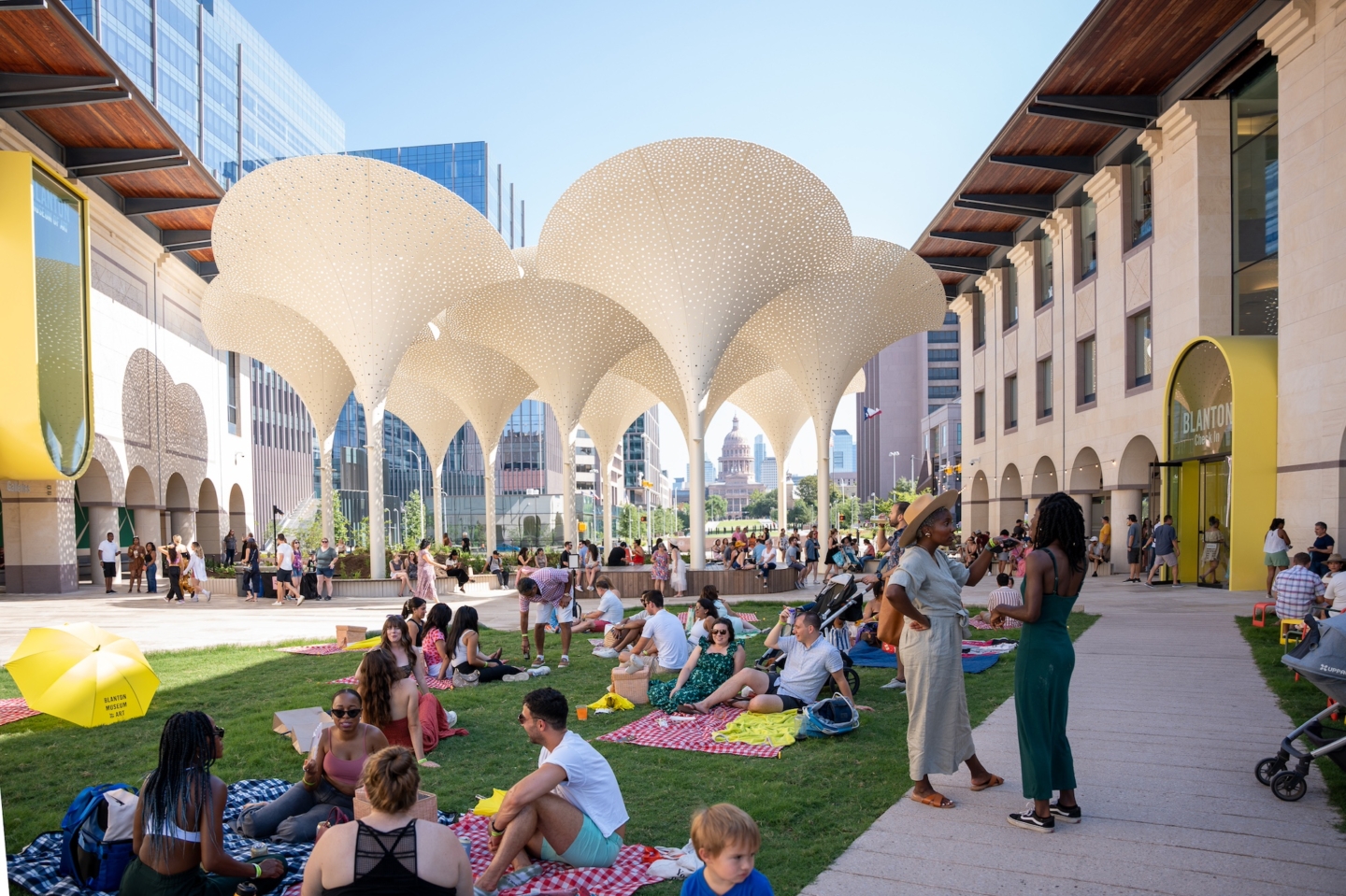 The image shows an outdoor area with a large, modern architectural canopy structure featuring multiple white, perforated, umbrella-like elements. The canopy provides shade for people gathered underneath. The area appears to be a public space, part of a museum. People are sitting on the grass and benches, engaging in various activities such as talking, relaxing, and enjoying the environment. The background includes tall buildings and a distant view of a domed structure, likely a capitol building, suggesting an urban setting. The scene is lively and vibrant, indicating a communal or social event.