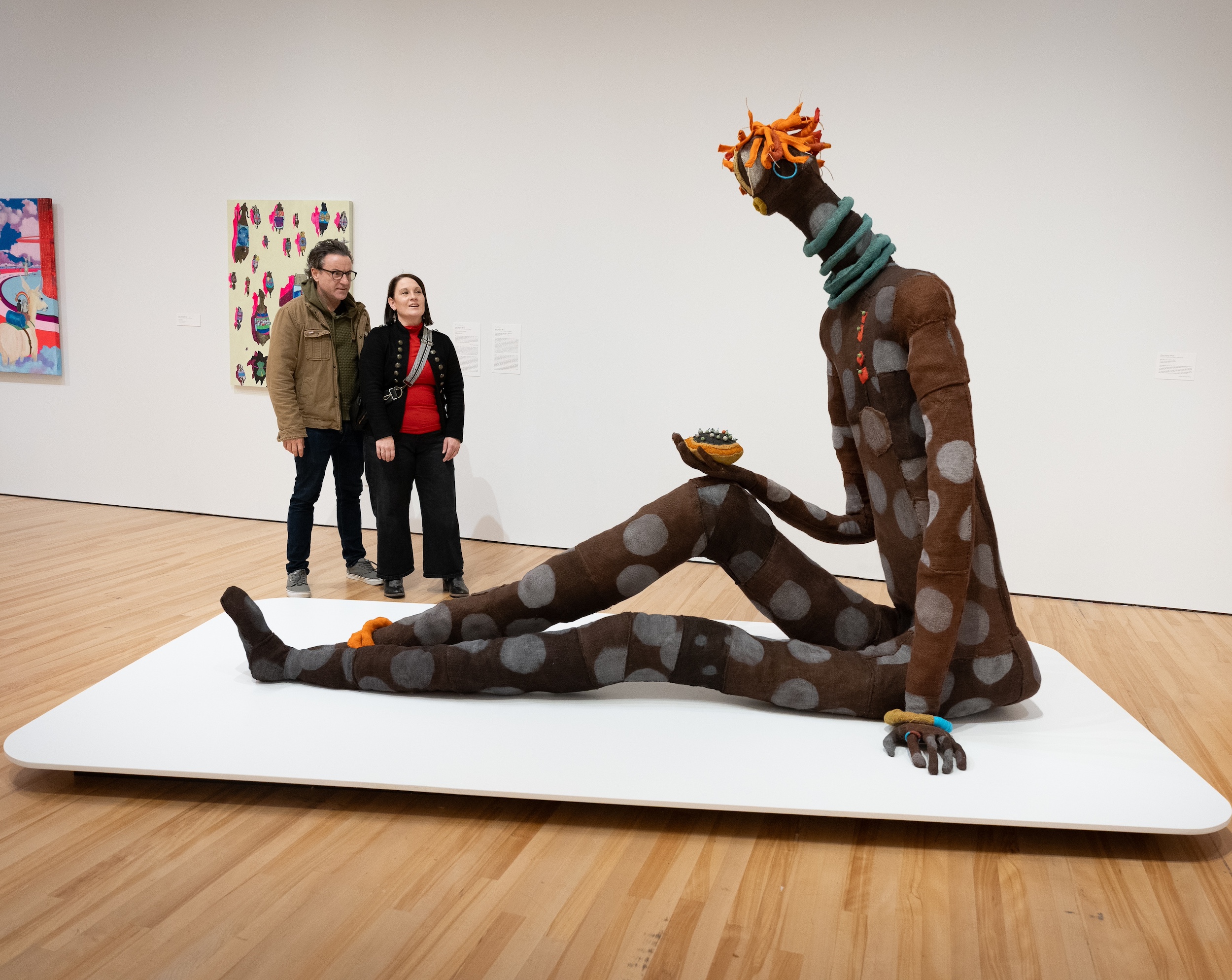 Two people looking at a large figure seated on a white plinth. It is extraordinarily large and made from burlap and stuffed with various materials, with large spots all over the body and red-coloured hair