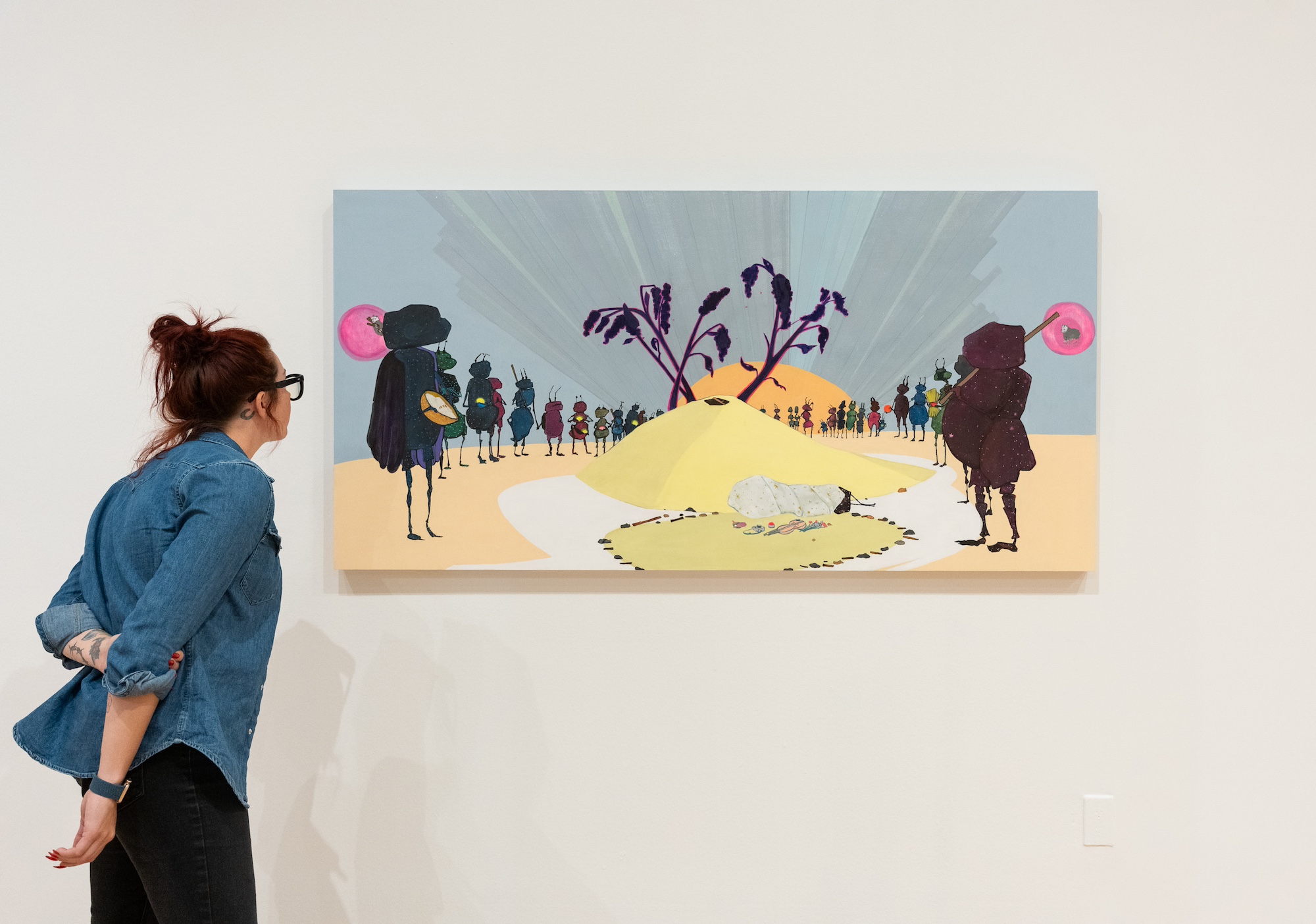 A person looking at a long, colorful painting on a wall featuring ant-like creatures gathered around a mound of earth with a plant sprouting out of it