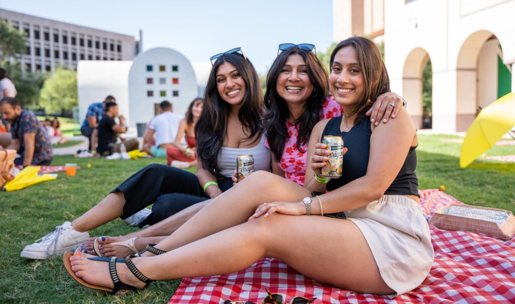 A mother with two daughters, smiling and sitting on a picnic blanket on the grounds of the Blanton Museum of art in Austin, Texas